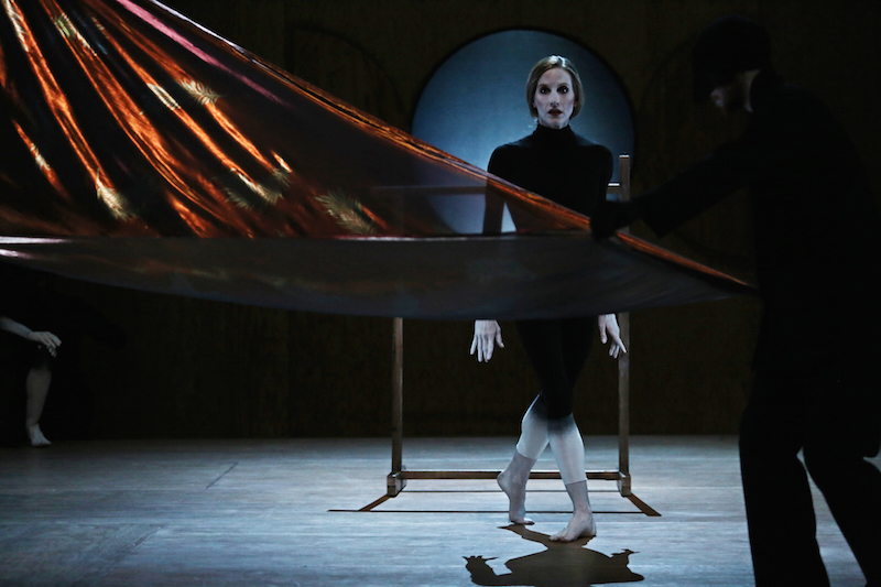 Wendy Whlean dressed in black stares at the audience as a cascade of fabric unfurls before her.